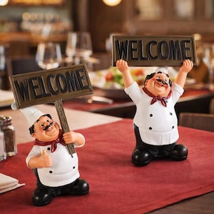 May include: Two chef figurines, one holding a "WELCOME" sign and the other with the sign raised above its head. The figurines are dressed in white chef coats, black pants, and red neckerchiefs. The signs are brown with black lettering.