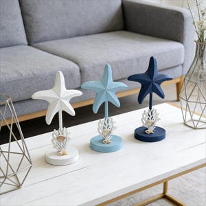 Könnte beinhalten: Drei dekorative Seestern-Skulpturen in Weiß, Hellblau und Marineblau. Jeder Seestern ist auf einem Ständer mit Korallen- und Muscheldetails montiert. Küsten-Wohndekoration.