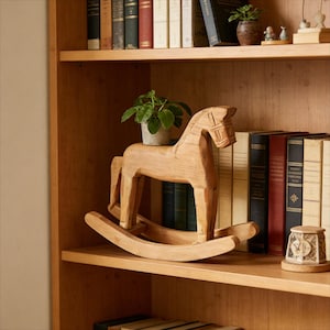 Pode incluir: Uma escultura de cavalo de baloiço de madeira com uma pequena planta em vaso nas costas está numa prateleira. O cavalo é castanho claro e tem um design esculpido. Livros e outros objetos decorativos estão nas prateleiras.