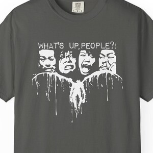 Puede incluir: Una camiseta de manga corta de color gris carbón con un estampado gráfico blanco de cuatro rostros con expresiones distorsionadas. Sobre los rostros, el texto "WHAT'S UP, PEOPLE?!" con un efecto de pintura que gotea hacia abajo.
