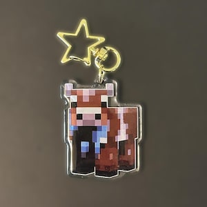Puede incluir: Un llavero acrílico transparente con un diseño pixelado de vaca marrón y blanca con una bufanda azul. El llavero tiene un dije dorado en forma de estrella y un anillo para sujetar llaves o accesorios.