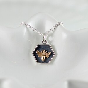 May include: A silver chain necklace with a hexagonal pendant featuring a gold bee design. The pendant is black with a silver border.