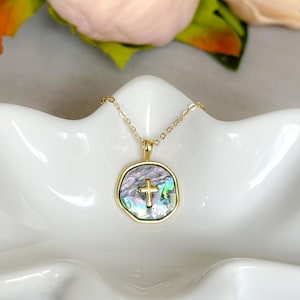 May include: A gold-toned necklace with a cross pendant featuring an iridescent abalone shell inlay. The necklace rests in a white, petal-shaped dish, with soft pink flowers in the background. The pendant's cross is centered within the shell.