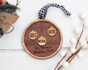 May include: A round wooden Christmas bauble with the names Greg, Katie, and Nora engraved on it. The bauble also has the text "The Woodles Christmas 2020" engraved on it.