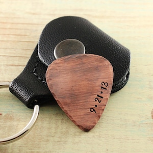 May include: A wooden guitar pick keychain with a black leather case and a silver key ring. The pick is engraved with the date "9.21.13".