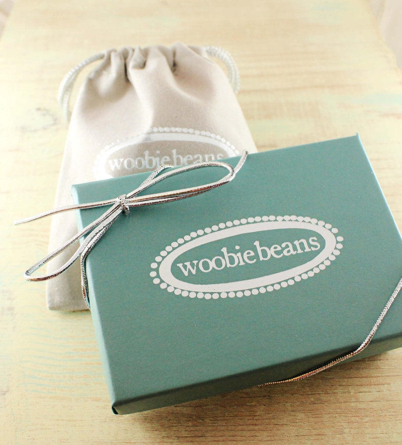May include: A teal gift box with a white oval label that says "woobiebeans" in a white font. The box is tied with a silver ribbon and is sitting on a light wood surface.