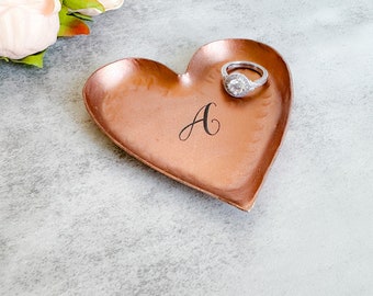 May include: A heart-shaped copper ring dish with a hammered texture. The dish has the letter "A" engraved on it and a silver ring with a large, clear stone is resting on the dish.