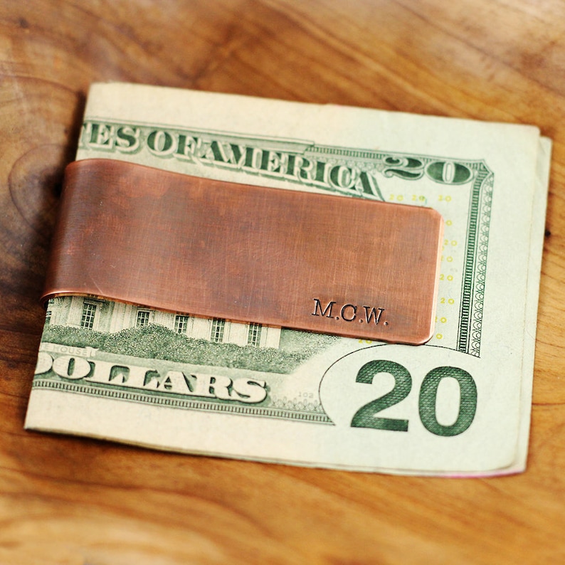 May include: A copper money clip with the initials "M.C.W." engraved on it. The clip is holding a twenty dollar bill.