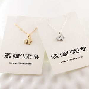 May include: Two gold and silver necklaces with bunny charms. The charms have the letters "E" and "S" engraved on them. The necklaces are on white cards with the text "Some bunny loves you." and the website address "www.woobiebeans.com".