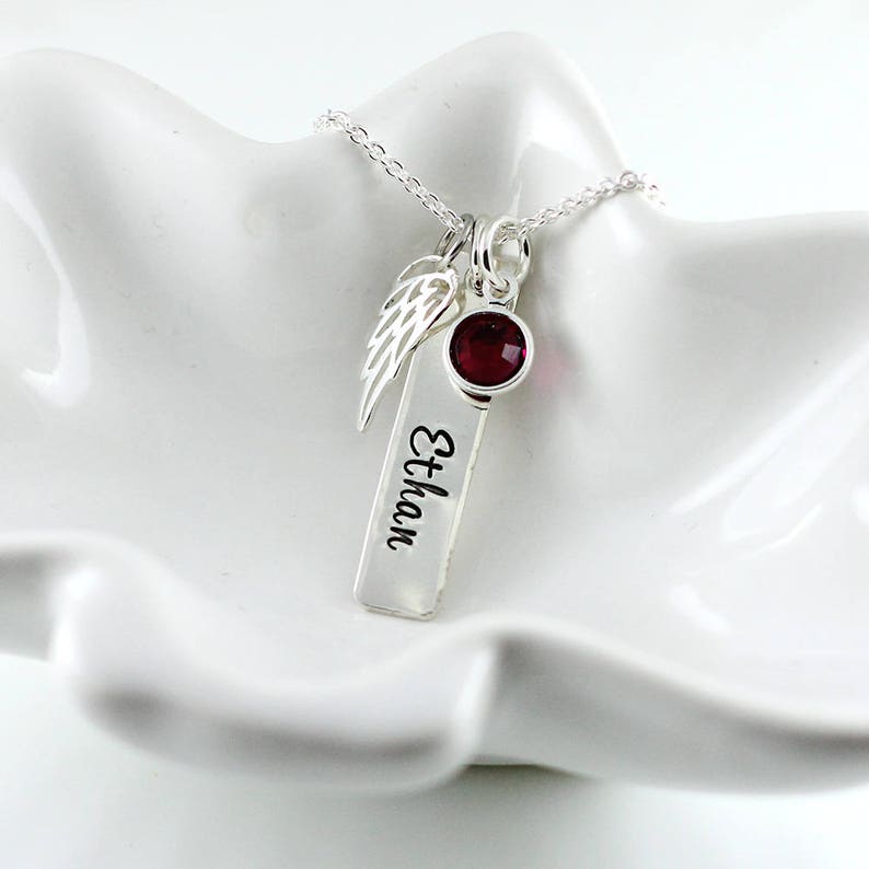 May include: Silver necklace with a small red gemstone, a silver angel wing charm, and a silver bar charm engraved with the name "Ethan".