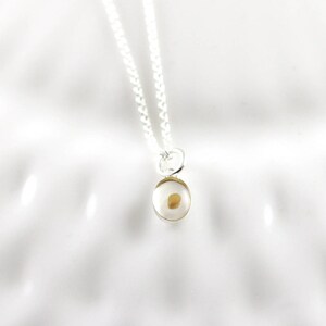 May include: A silver necklace with a small, clear pendant containing a mustard seed. The pendant is oval-shaped with a gold-colored rim. The chain is a delicate silver link.
