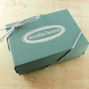 May include: A teal blue gift box with a silver ribbon tied in a bow. The box has the words "woobie beans" printed on the top in white.