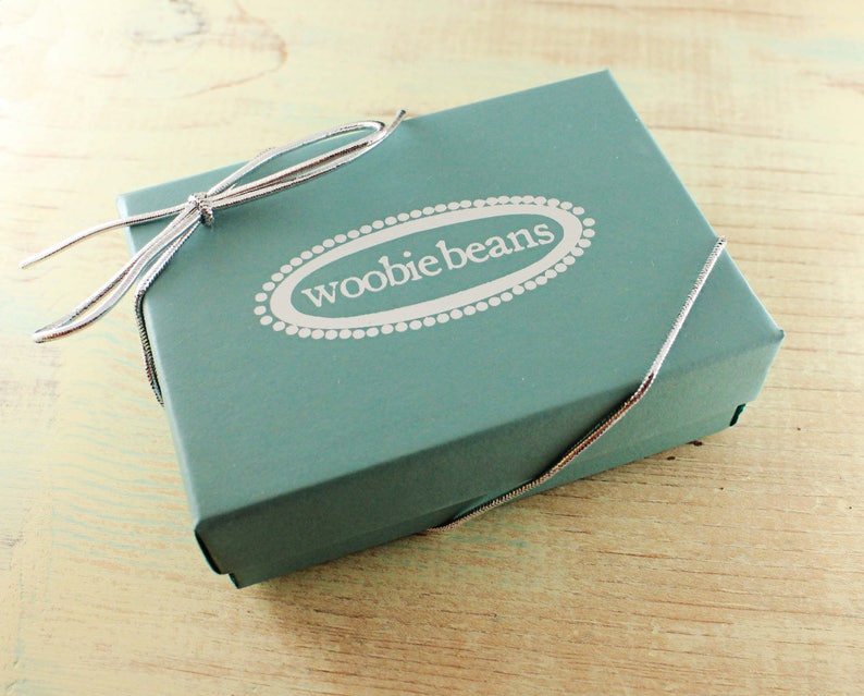 May include: A teal blue gift box with a silver ribbon tied in a bow. The box has the words "woobie beans" printed on the top in white.