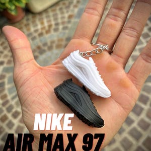 Può includere: Miniature di scarpe da ginnastica Nike Air Max 97 bianche e nere attaccate a un portachiavi. Le scarpe da ginnastica sono repliche dettagliate dell'iconico design delle scarpe. Il testo "NIKE AIR MAX 97" è visualizzato in bianco e rosso.