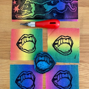 Può includere: Una collezione di linoleografie e strumenti. Le stampe presentano una silhouette nera di un animale che salta con stelle su uno sfondo arcobaleno e bocche aperte con zanne su uno sfondo arcobaleno. È presente anche uno strumento di linoleografia rosso.