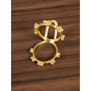 Könnte beinhalten: Ein goldfarbener Ring mit einem einzigartigen Design. Der Ring hat zwei verbundene kreisförmige Bänder, eines größer als das andere, mit einer Reihe kleiner, runder Verzierungen. Der obere Teil hat einen dekorativen Bogen und vertikale Stäbe. Der Ring ist vor einem dunklen Holzmaserungshintergrund platziert.