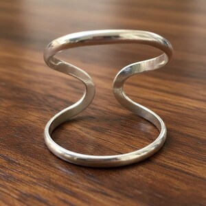 Puede incluir: Un anillo plateado con un diseño único. El anillo presenta un diseño curvo y abierto con dos bandas paralelas conectadas por un elemento central ondulado. El anillo está hecho de un material metálico brillante.
