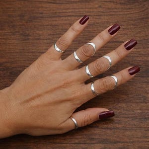 May include: A hand displaying several silver rings of varying designs. The rings include simple bands and open-ended crescent shapes. The nails are painted a dark burgundy color, set against a dark wood background.