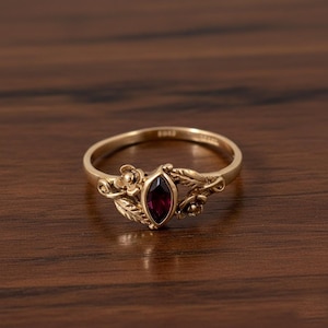 Op de afbeelding: Een goudkleurige ring met een dieprode, marquise-geslepen edelsteen. De ring heeft bloemen- en blad details op de band, met kleine bloemen en bladeren aan weerszijden van de edelsteen. De ring is geplaatst op een donker houten oppervlak.