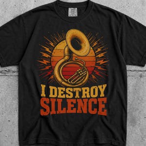 Peut inclure: Un t-shirt noir à manches courtes orné d'un tuba doré centré devant un cercle aux couleurs du coucher de soleil avec des lignes rayonnantes. Sous l'instrument, le texte I DESTROY SILENCE est écrit en lettres capitales orange-rouge.