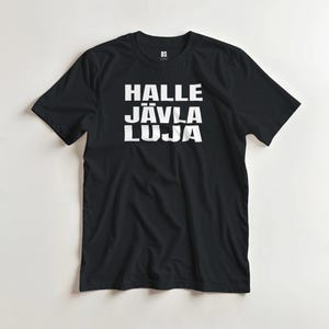 Puede incluir: Camiseta negra con el texto blanco "HALLE JÄVLA LUJA" apilado verticalmente. La camiseta tiene mangas cortas y cuello redondo. El logotipo de la marca "BS" está en la esquina superior derecha.