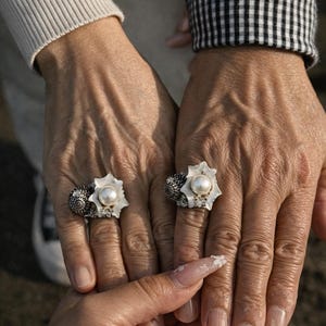 Puede incluir: Dos manos que muestran anillos con un centro de perla blanca y una banda plateada decorativa. Los anillos tienen un diseño floral alrededor de la perla. Las manos están frente a un fondo neutro.