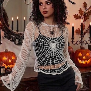 Puede incluir: Una blusa de ganchillo blanca de manga larga con un patrón de telaraña en la parte delantera y las mangas. La prenda se lleva sobre una camiseta negra y se combina con una falda negra. La blusa tiene un cuello redondo y mangas holgadas.