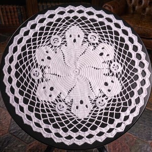 May include: A round white crochet doily featuring a central pattern of six ghost figures arranged in a circular formation, interspersed with small circular motifs, all surrounded by an intricate openwork lace border with a scalloped edge.