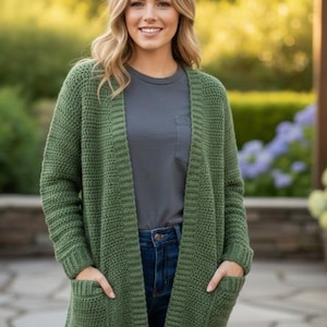 May include: A green knit cardigan with two front pockets. The open-front sweater has a textured pattern and ribbed cuffs. The person in the image is wearing a gray t-shirt and blue jeans. The background is blurred with greenery.