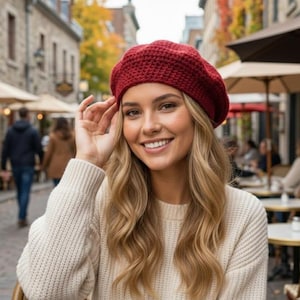 May include: A burgundy red crochet beret. The beret is worn with a cream-colored sweater. The background shows a European street scene with outdoor cafes and people walking. The beret is a stylish accessory.
