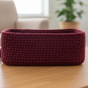 Puede incluir: Una cesta rectangular de ganchillo burdeos se encuentra sobre una mesa de madera. Una madeja de hilo a juego descansa a su lado. La cesta está hecha a mano y tiene un aspecto texturizado, adecuada para guardar o decorar.