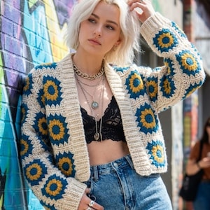 Può includere: Giacca all'uncinetto color crema con motivi floreali blu e gialli. La giacca è indossata sopra un top in pizzo nero e abbinata a jeans blu. Il design della giacca presenta motivi quadrati con un motivo a forma di girasole.