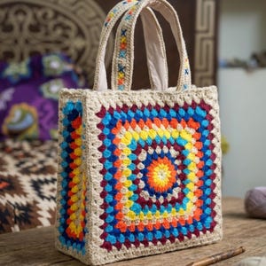 May include: A square crochet tote bag featuring a vibrant granny square pattern in shades of orange, yellow, blue, and maroon on a beige background. The bag has two matching beige crochet handles with colorful cross-stitch embroidery details.