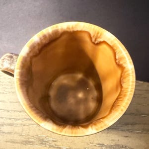 May include: A ceramic mug with a brown interior and a tan and brown rim. The mug has a handle and a glossy finish. The interior of the mug is a darker brown color.