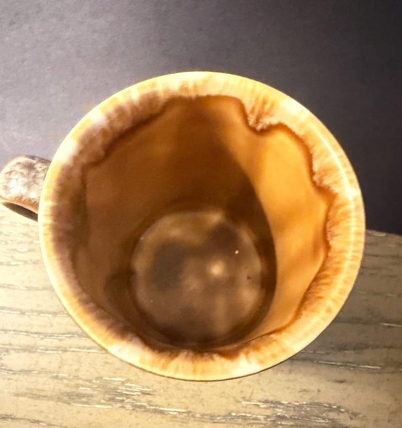 May include: A ceramic mug with a brown interior and a tan and brown rim. The mug has a handle and a glossy finish. The interior of the mug is a darker brown color.