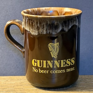 May include: A brown ceramic Guinness mug with a gold harp logo and the text "GUINNESS No beer comes near." The mug has a dark brown exterior and a lighter brown rim with a white and brown drip design.