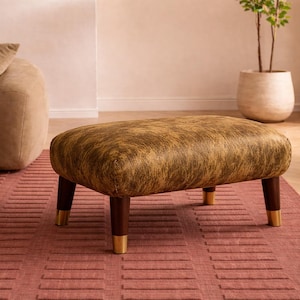 Pode incluir: Um pufe estofado com um tampo com padrão castanho e dourado e pernas de madeira escura com pontas douradas. O pufe retangular está sobre um tapete vermelho numa sala de estar.