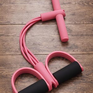 Può includere: Attrezzo per esercizi con fasce di resistenza rosa e manici in schiuma neri. L'attrezzo per il fitness ha una maniglia superiore a forma di T e due anelli per i piedi. Le fasce sono curve.