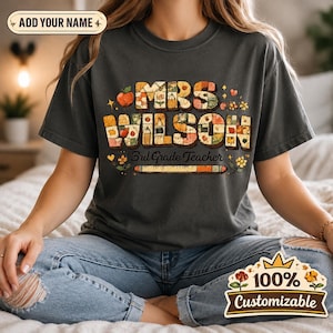 以下が含まれることがあります： チャコールグレーの半袖Tシャツ。学校をテーマにした模様で埋め尽くされたブロック体の「MRS WILSON」という文字、筆記体の「3rd Grade Teacher」、リンゴ、花、鉛筆のイラストが描かれたグラフィックプリントが施されています。