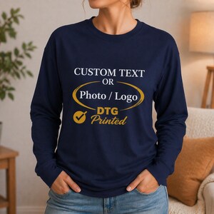 May include: Navy blue long-sleeve shirt with the text "CUSTOM TEXT OR Photo / Logo DTG Printed" in white and gold. The shirt has a crew neck and is made of a soft material.