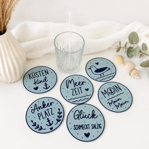 Può includere: Un set di sottobicchieri blu chiaro con disegni a tema nautico e testo in tedesco, tra cui ancore, gabbiani e frasi come "Meer Zeit" e "Glück schmeckt salzig". Sono presenti anche un bicchiere trasparente e una cannuccia.