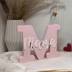 May include: A pink wooden letter "M" with the word "Mama" in white script. A small glass vial with a cork stopper is inserted into the letter. The decor is on a wooden slice and white cloth.