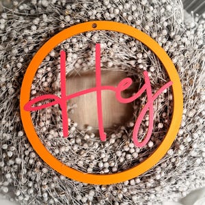 Könnte beinhalten: Ein dekorativer Kranz mit einem kreisförmigen orangefarbenen Rahmen und dem Wort "Hey" in rosa Schrift. Der Kranz besteht aus getrockneten weißen Beeren und Stielen und erzeugt ein natürliches, strukturiertes Aussehen. Geeignet für die Inneneinrichtung.