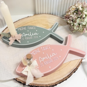 Pode incluir: Duas lembrancinhas de batismo em forma de peixe, uma verde sálvia e outra rosa, cada uma com o texto "MEINE TAUFE" e o nome "Amilia" com a data "20. Februar 2026". O peixe verde segura uma vela e o rosa uma garrafa pequena.