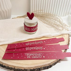 Könnte beinhalten: Ein kleines Glas-Kerzenglas mit rotem herzförmigem Deckel und rotem Band mit der Aufschrift "Beste Freundin". Drei rote Lesezeichen mit weißem Text und Punkten liegen auf einer Holzoberfläche. Auf den Lesezeichen steht "Herzensfreundin", "Seelenfreundin" und "Freundschaft, die leuchtet".