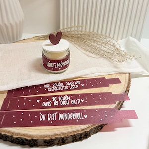 Può includere: Una piccola candela in barattolo di vetro con un coperchio a forma di cuore rosso e la scritta "HerzMensch" sull'etichetta. Tre etichette bordeaux con testo bianco e pois sono disposte su una fetta di legno.