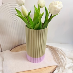 Op de afbeelding: Een decoratieve vaas met een geribbeld ontwerp in olijfgroen en lavendel, met witte tulpen met groene bladeren. De vaas staat op een houten schijf met een witte doek.