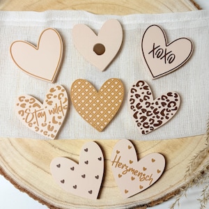 May include: A collection of heart-shaped decorations in various designs and colors. Some hearts feature leopard print, while others have text like "Stay Wild" and "XOXO." The hearts are arranged on a wooden surface.