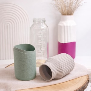 May include: A still life arrangement featuring decorative vases and a clear glass bottle. The vases are in shades of green, white, and pink. One vase is textured, and another has a ribbed design. The bottle has text on it.