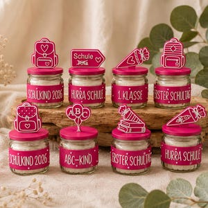 May include: Eight small glass jars with white wax, each featuring a bright pink lid and a matching pink band around the center. The lids have various pink decorative toppers, and the bands display white text including Schulkind 2026, Hurra Schule, 1. Klasse, Erster Schultag, and ABC-Kind.
